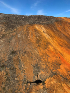 Aerial view of a rusty-orange oxidised hillside in rocky Arctic terrain, with a helicopter shadow