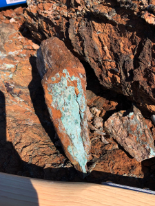 Close-up of rust-coloured oxidised rock with pale green secondary mineral coating and a hammer handle