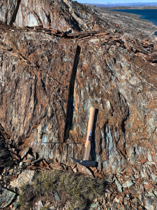 Close-up of a channel-sample cut in fractured rock with a geological hammer, fjord and mountains behind