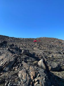 Red helicopter flying over a steep rocky Arctic slope under a clear blue sky