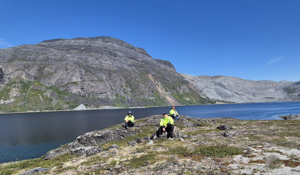 Three people in high-visibility jackets sitting on a rocky point beside a lake in South Greenland